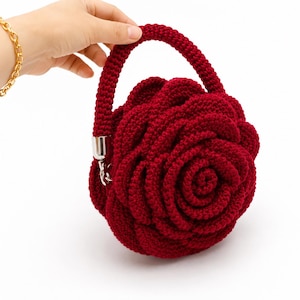 May include: A deep red, crocheted handbag in the shape of a rose. It has a rounded handle and a silver-coloured clasp. The rose design is detailed with spiralling petals, creating a three-dimensional effect. The bag is held by a hand.