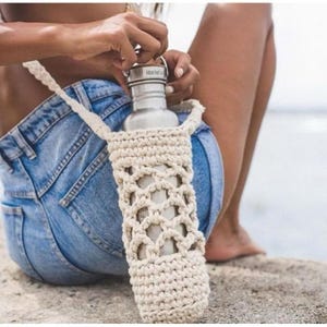 May include: A stainless steel water bottle in a cream-colored macrame carrier with a shoulder strap. The carrier has an open, woven design. The bottle is being opened by a person wearing denim shorts.