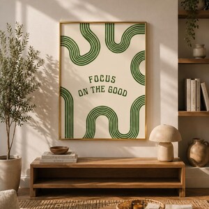 Könnte beinhalten: Ein rechteckiger Wandkunstdruck in einem goldenen Rahmen mit cremefarbenem Hintergrund, dunkelgrünen wellenförmigen Linienmustern und dem Text "FOCUS ON THE GOOD" in einer dunkelgrünen serifenlosen Schrift.