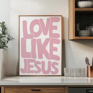Könnte beinhalten: Gerahmter Kunstdruck mit den Worten "LOVE LIKE JESUS" in großen, rosa Buchstaben im Blasenstil auf weißem Hintergrund. Der Rahmen ist hellbraun. Das Kunstwerk wird in einer Küchenumgebung ausgestellt.