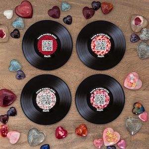 May include: Four black vinyl record coasters with custom labels featuring a QR code, the text "YOUR NAMES HERE" and "DATE". Decorative heart-shaped objects in various colours surround the coasters.