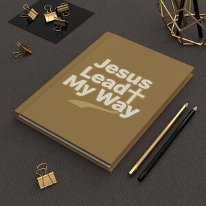 以下が含まれることがあります： 「Jesus Lead My Way」という白い文字と十字架のシンボルが描かれた、タンカラーのハードカバーノート。ノートの横にはゴールドとブラックの鉛筆が2本置かれ、近くにはゴールドの金属製バインダークリップがいくつか散らばっています。