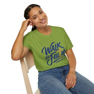 Puede incluir: Una camiseta verde lima con las palabras "Walk by Faith" en escritura azul y una cruz dorada. El texto "2 Corintios 5:7" está debajo. La persona lleva la camiseta y está sentada en una silla de madera.