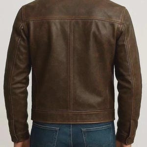 May include: A brown leather jacket with a button-down collar and long sleeves. The jacket has a classic design with a tailored fit and visible stitching details. The model is wearing blue jeans.