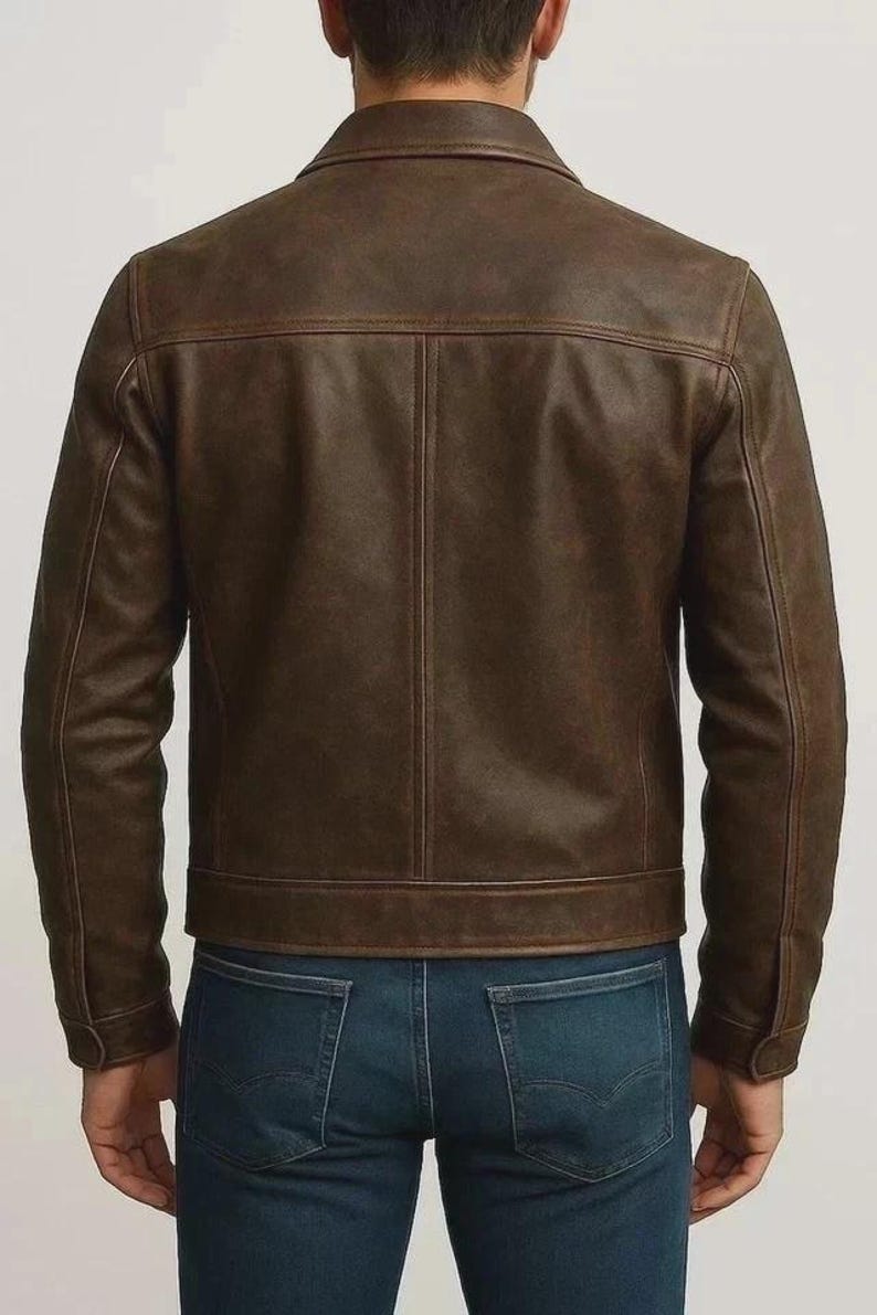 May include: A brown leather jacket with a button-down collar and long sleeves. The jacket has a classic design with a tailored fit and visible stitching details. The model is wearing blue jeans.