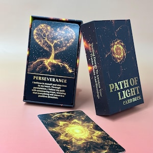 May include: A card deck with a navy blue box and cards. The box reads "Path of Light Card Deck" in gold lettering. The cards feature gold and black designs with the word "Perseverance" and a description. The deck is on a pink surface.