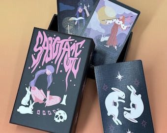 Puede incluir: Una caja negra de cartas del tarot con la palabra "SABOTAGE YOU" en letras rosas. La caja presenta ilustraciones de una mujer, conejos y una calavera. Se ven varias cartas del tarot con ilustraciones de figuras y conejos.