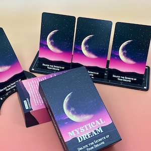 May include: A Mystical Dream card deck featuring a crescent moon design against a starry night sky with pink and purple tones. The box and cards display the text "Unlock the Secrets of Your Dreams". The deck is presented on a pink surface.