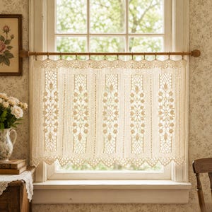 May include: A cream-colored cafe curtain featuring an intricate crochet lace pattern with vertical floral motifs and a scalloped bottom edge. The curtain is suspended from a wooden rod with matching rings, covering the lower half of a window.