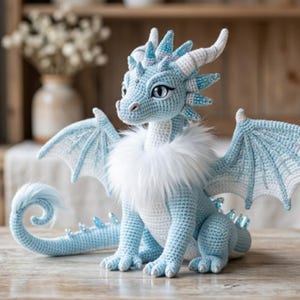 May include: A crocheted dragon figurine in shades of light blue and white. The dragon has detailed wings, horns, and a fluffy white collar. The dragon is sitting on a wooden surface.