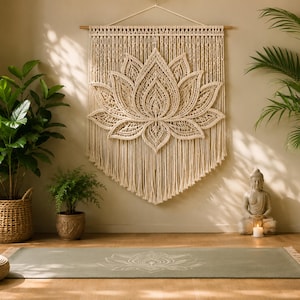 Può includere: Un grande arazzo da parete in macramè color bianco sporco con un intricato disegno a fiore di loto appeso a un bastone di legno. Il pezzo è composto da corda di cotone annodata con un bordo inferiore sfrangiato e motivi geometrici.