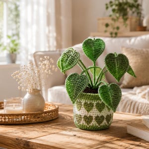 May include: A crocheted plant with heart-shaped green leaves potted in a cylindrical container featuring a repeating pattern of green and white hearts. The plant is crafted with textured yarn stitches, showcasing a vibrant green color palette.