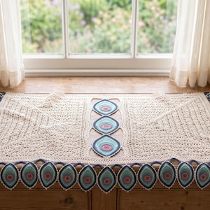 May include: A rectangular cream-colored crochet table runner featuring an intricate openwork lace pattern. The runner is decorated with a central vertical row of three and a bottom horizontal row of ten teardrop-shaped motifs in shades of blue, teal, and pink.