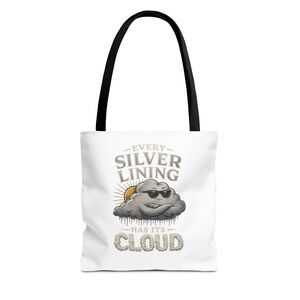 May include: White tote bag with black handles featuring the text "EVERY SILVER LINING HAS ITS CLOUD". The design includes a cartoon cloud wearing sunglasses with a sun in the background. The bag is made of a durable material.