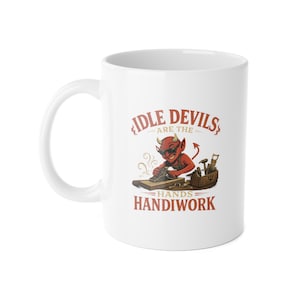 May include: White ceramic mug featuring a cartoon devil with sunglasses, horns, and a tail, working with tools. The text on the mug reads "Idle Devils are the Hands Handiwork" in red lettering. The mug has a white handle.