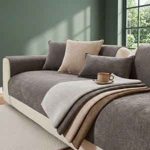 May include: A sofa covered in a textured dark gray fabric with a cream-colored frame. The sofa features a square beige cushion, a rectangular dark gray cushion, and a layered throw blanket in shades of gray and beige, topped with a small ceramic mug.