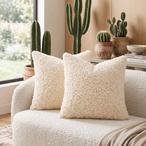 May include: Two square throw pillows made of cream-colored, textured sherpa fabric. The pillows are positioned on a matching cream-colored sofa, featuring a thick, curly, and plush pile surface that provides a soft and tactile appearance.