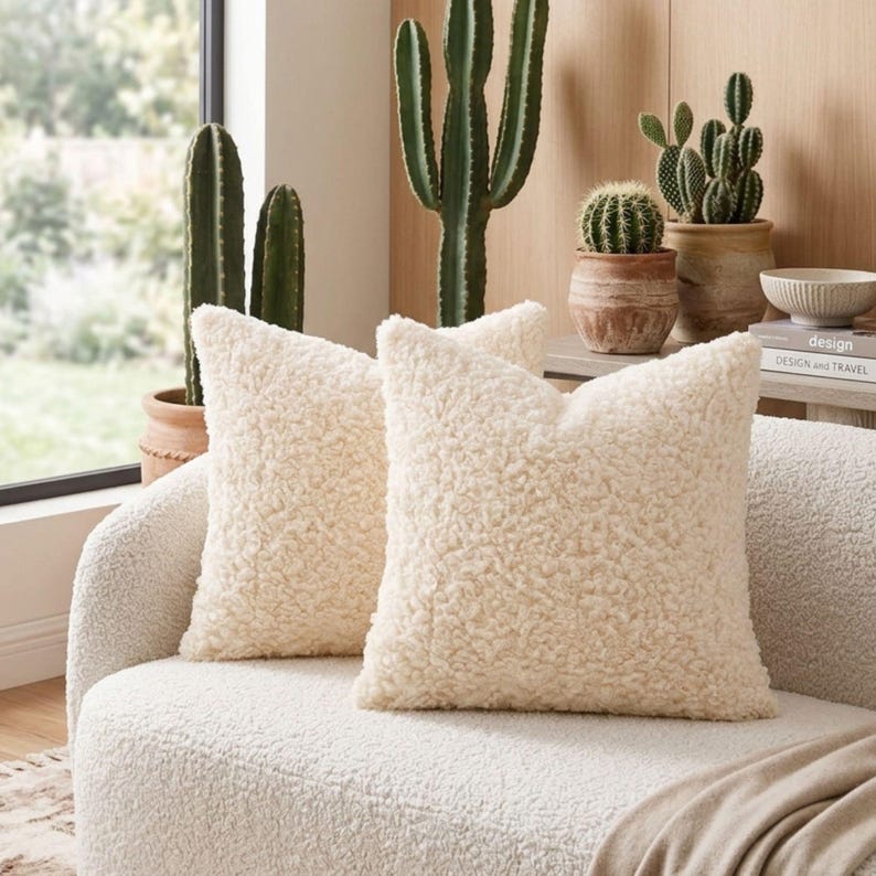May include: Two square throw pillows made of cream-colored, textured sherpa fabric. The pillows are positioned on a matching cream-colored sofa, featuring a thick, curly, and plush pile surface that provides a soft and tactile appearance.
