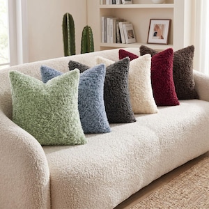 May include: A collection of seven square throw pillows with a textured, curly faux-shearling fabric. The pillows are arranged in a row on a sofa and feature a variety of colors including sage green, dusty blue, charcoal gray, cream, and deep burgundy.