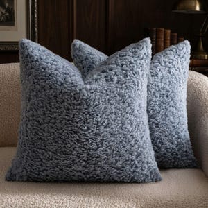 May include: Two square throw pillows covered in a dusty blue, high-pile sherpa fabric. The material features a dense, curly, and textured surface that creates a soft, voluminous appearance. The pillows are positioned upright, showcasing the uniform, shaggy texture across the entire front and side surfaces of the square cushions.