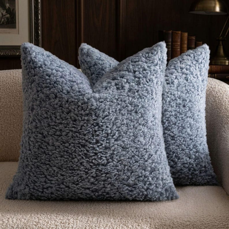 May include: Two square throw pillows covered in a dusty blue, high-pile sherpa fabric. The material features a dense, curly, and textured surface that creates a soft, voluminous appearance. The pillows are positioned upright, showcasing the uniform, shaggy texture across the entire front and side surfaces of the square cushions.