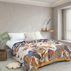 Könnte beinhalten: Ein Bett mit einer cremefarbenen Tagesdecke mit einem lebendigen botanischen Druck in Senfgelb, Dunkelblau und Magenta. Das Bett ist mit zwei einfarbigen weißen Kissen, einem beigen Kissen und einem weißen Kissen mit gelben Blumenmustern dekoriert.