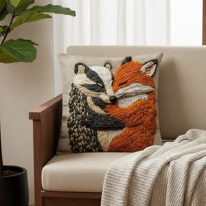 Puede incluir: Cojín decorativo bordado con un tejón y un zorro abrazándose. El cojín tiene un fondo beige y los animales están representados en tonos naranja, negro, blanco y marrón. El cojín está colocado sobre una silla beige con una manta gris.