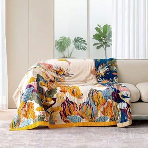 May include: A rectangular throw blanket featuring a cream-coloured base with a vibrant botanical print of stylised flowers and leaves in shades of mustard yellow, deep blue, purple, and teal, finished with a solid mustard yellow border trim.