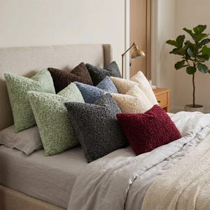 May include: A collection of square throw pillows with a textured, curly fleece-like surface. The pillows are arranged in various colors including sage green, dark brown, charcoal gray, light blue, cream, and deep burgundy on a bed.