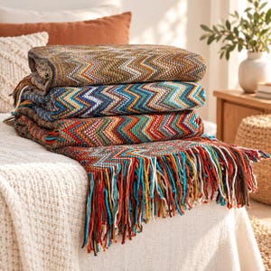 May include: A stack of three folded knit blankets featuring a multi-coloured chevron pattern in shades of brown, blue, orange, and white. The bottom blanket has long, dangling fringe tassels in matching colours along the edge of the fabric.