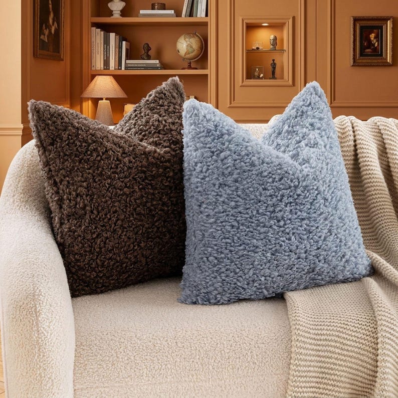 May include: Two square throw pillows with a thick, curly, sherpa-like textured fabric. One pillow is dark brown and the other is a light, dusty blue. They are positioned side-by-side on a cream-colored sofa with a matching knit throw blanket.