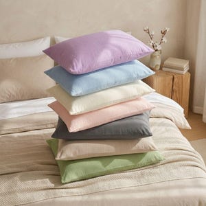 May include: A vertical stack of seven rectangular pillows in various solid colors including lavender, light blue, cream, soft pink, charcoal gray, beige, and sage green. Each pillow features a textured fabric surface and a simple rectangular shape.