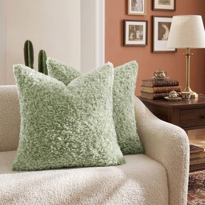 May include: Two square throw pillows featuring a textured, sage green faux shearling or sherpa fabric. The pillows have a dense, curly pile surface and are positioned on a cream-colored textured sofa, showcasing a soft and plush appearance.