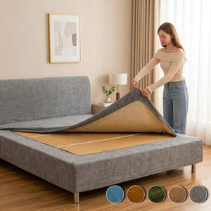 Op de afbeelding: Een grijs gestoffeerd bedframe met een bijpassend grijs hoeslaken dat wordt opgetild om een beige geweven mat eronder te onthullen. Het bed heeft een hoog, rechthoekig hoofdeinde en een lage basis met korte, metalen cilindrische poten.