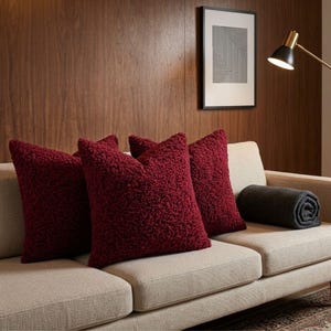 May include: Three square throw pillows in a deep burgundy color with a textured, curly sherpa-style fabric. The pillows are arranged on a light beige sofa alongside a rolled-up dark gray blanket, set against a dark wood paneled wall.