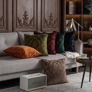 Può includere: Una collezione di cuscini quadrati con intricati motivi damascati nelle tonalità arancione, verde oliva, rosso intenso, verde scuro e nero, disposti su un divano grigio chiaro accanto a un altoparlante Marshall bianco sul pavimento.
