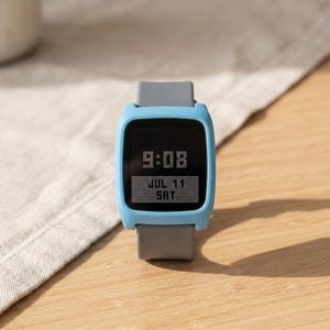 Puede incluir: Un reloj de pulsera digital con un marco cuadrado azul claro y una correa de silicona gris lisa. La pantalla negra muestra la hora 9:08 en una fuente pixelada, con la fecha JUL 11 y el día SAT en un recuadro blanco debajo.
