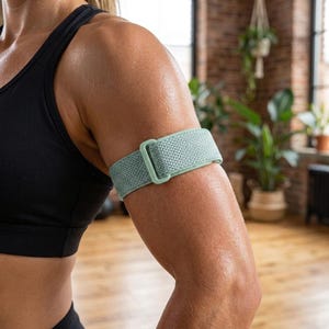 May include: A sage green elastic arm band featuring a textured woven fabric surface and a matching rectangular plastic buckle. The band is designed to wrap around the upper arm, providing a secure fit with its adjustable loop and hook closure.