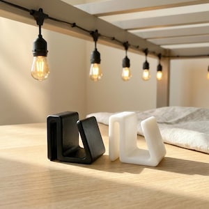 May include: Two U-shaped clips, one matte black and one matte white, featuring a sturdy, thick-walled design with a flat base and a curved top edge. These functional clips are positioned side-by-side, showcasing their smooth, solid construction.