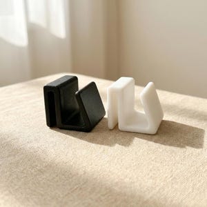 May include: Two U-shaped clips, one in matte black and one in matte white, featuring a smooth, solid construction with a distinct channel design. These two small, functional objects are positioned side-by-side, showcasing their uniform shape.