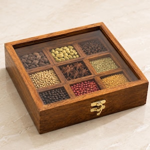May include: A square wooden spice box with a clear glass lid and a gold-toned metal latch. The box is divided into nine individual compartments, each containing a different variety of whole spices, including star anise, peppercorns, and seeds.