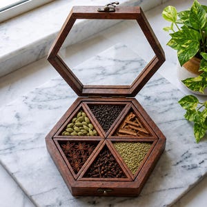 May include: A hexagonal wooden spice box with a hinged glass lid, featuring six triangular compartments filled with various whole spices including green cardamom pods, black peppercorns, cinnamon sticks, star anise, cloves, and fennel seeds.