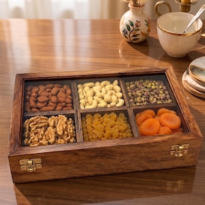 May include: A rectangular wooden storage box with a clear glass lid and two gold-toned metal latches on the front. The interior is divided into six square compartments, each filled with a different variety of dried fruits and assorted nuts.