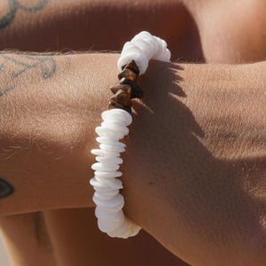Könnte beinhalten: Ein Armband bestehend aus einer Reihe kleiner, flacher, kreisförmiger weißer Scheibenperlen in einer durchgehenden Schlaufe, akzentuiert durch eine Gruppe unregelmäßiger, dunkelbrauner, texturierter steinartiger Perlen am oberen Rand.