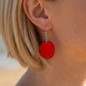Könnte beinhalten: Ein kreisförmiger roter Ohrring mit einer glatten, einfarbigen Scheibe, die an einem silberfarbenen Metallhaken hängt. Der Ohrring hängt vertikal und zeigt einen leuchtenden, gesättigten Rotton mit einer polierten Oberfläche.
