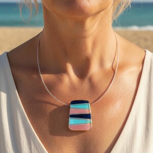 Könnte beinhalten: Halskette mit rechteckigem Anhänger und silberner Kette. Der Anhänger hat ein gestreiftes Design in Rosa, Hellblau und Marineblau. Die Halskette wird von einer Person mit blonden Haaren getragen, mit einem Strand im Hintergrund.