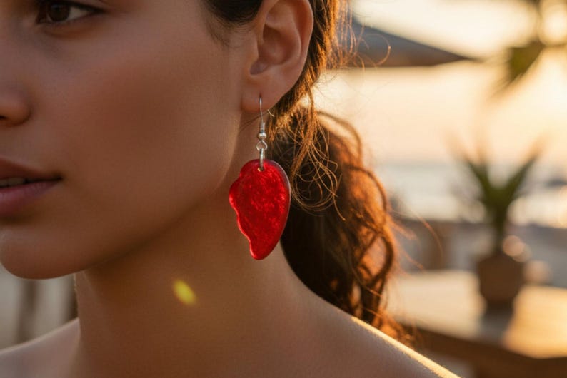 May include: A red, leaf-shaped earring hangs from a silver hook. The earring has a textured surface and is worn by a person with dark hair. The background is blurred, with warm sunlight.