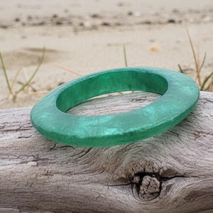 May include: A jade-green, circular bracelet sits on a piece of driftwood. The bracelet has a textured surface and a smooth, rounded shape. The background is a sandy beach with sparse vegetation.