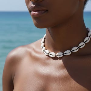Può includere: Primo piano di una persona che indossa una collana fatta di conchiglie bianche con dettagli neri. La collana è infilata con uno spago nero. Lo sfondo è una vista sfocata dell'oceano e del cielo.