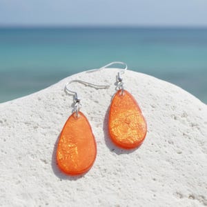 Op de afbeelding: Een paar oranje druppelvormige oorbellen met zilveren haken. De oorbellen hebben een glinsterend, getextureerd uiterlijk en worden gepresenteerd op een wit, getextureerd oppervlak met een vage oceaanachtergrond.